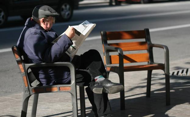 Un jubilado lee un periódico sentado en un banco de una calle. 