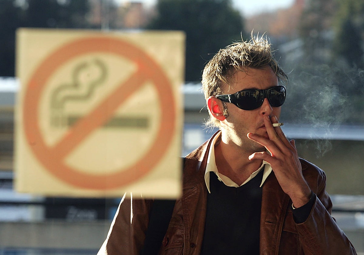 Un hombre fumando en frente de un cartel de «Prohibido fumar».