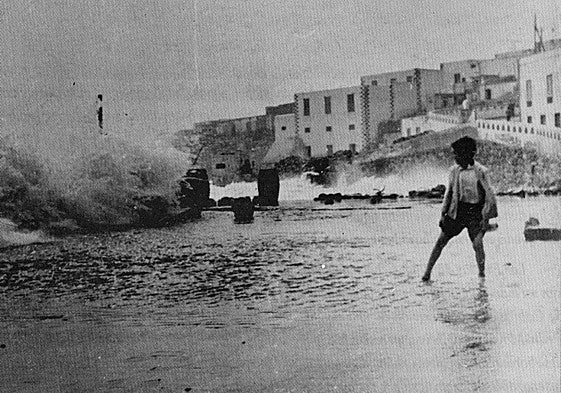 Un niño mira cómo una ola rompe en el muelle chico, con Puerto de Cabras al fondo.