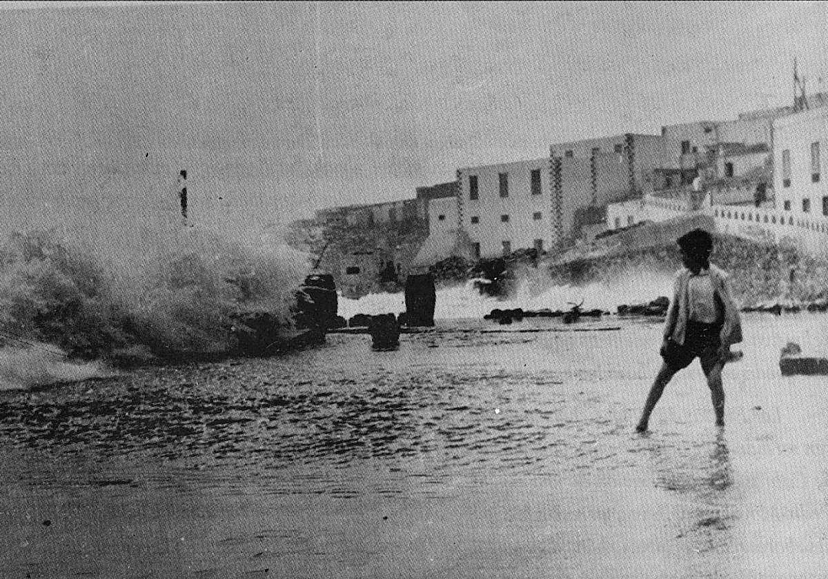 Un niño mira cómo una ola rompe en el muelle chico, con Puerto de Cabras al fondo.