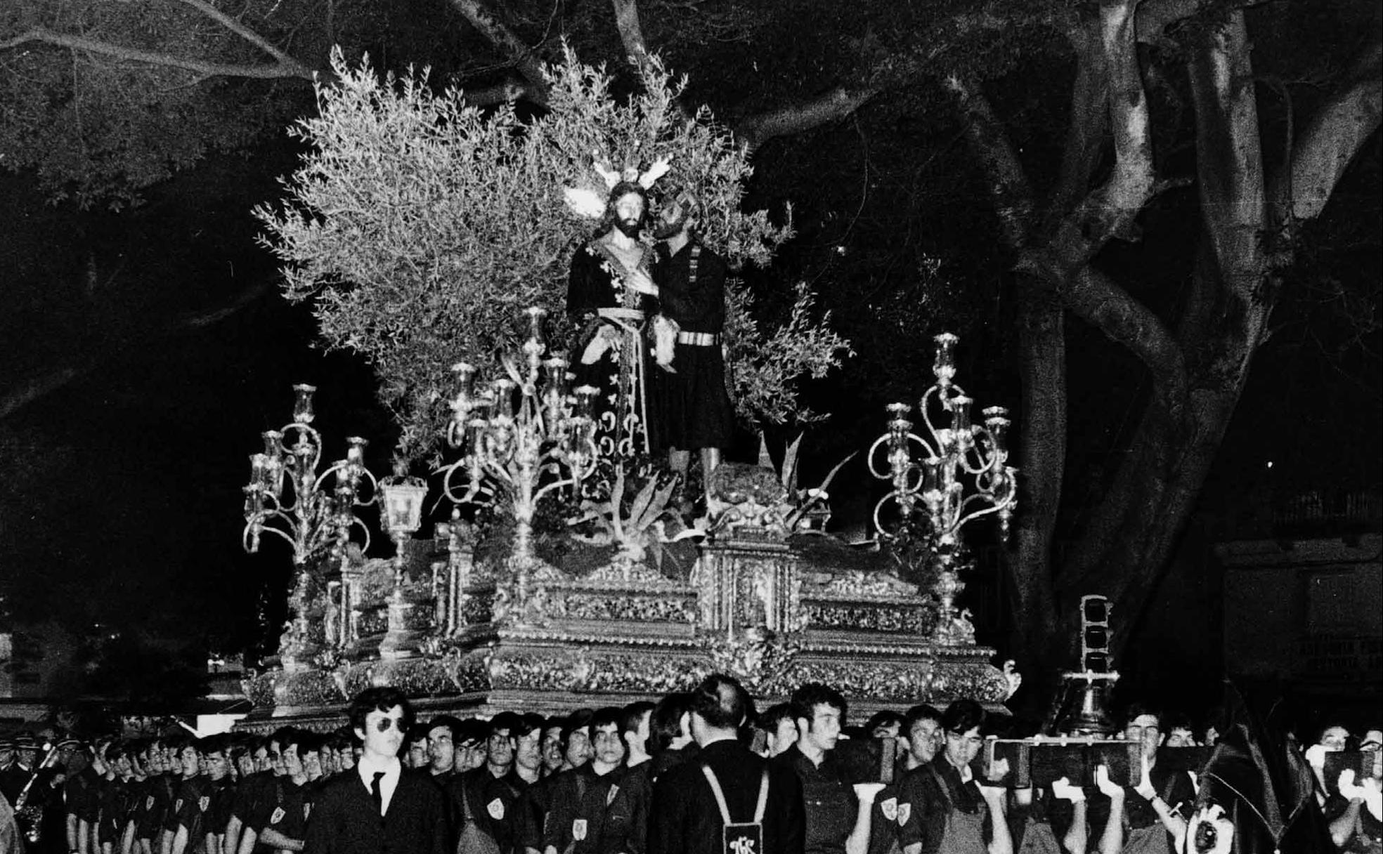 1970. El trono del Cristo, en la Alameda Principal, portado por alumnos de la Escuela Francisco Franco, ataviados con el característico mono de trabajo. 