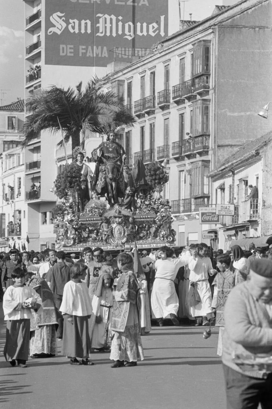 El trono de Jesús a su Entrada en Jerusalén avanza por el pasillo de Santa Isabel mientras empieza a caer la tarde. Un pequeño grupo de monaguillos precede a las andas en las que llama la atención la postura de uno de los cabezas de varal, metiendo el hombro de espaldas al sentido de la marcha.