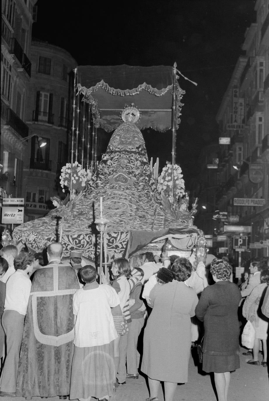 Curiosa fotografía de la trasera del trono de la Virgen del Gran Perdón en 1972, seguido por un sacerdote acompañado por monaguillos. Los hombres de trono han levantado el manto para que les entre algo de aire.