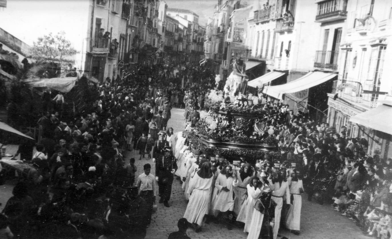 En el año 1954, la lluvia impidió los desfiles procesionales del Lunes Santo, por lo que las cofradías de esta jornada salieron a primeras horas de la tarde del día siguiente. En la fotografía se aprecia el paso del Señor de la Columna ante la Tribuna de los Pobres en una abarrotada calle Carretería.