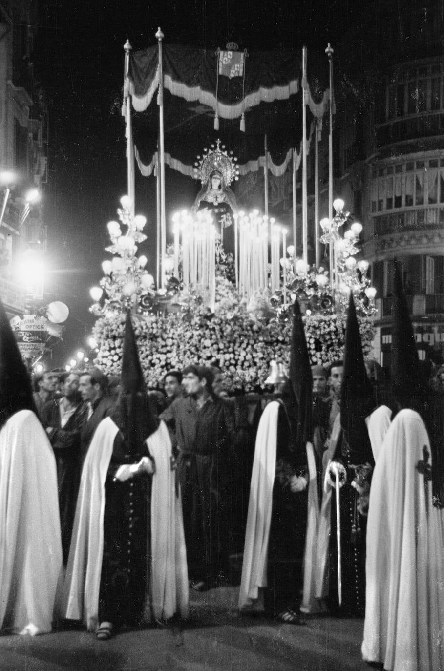 La Virgen del Amor procesionó durante varios años sobre cajillos en los que, junto a algunas tallas, predominaban las flores para suplir lo que debería ser madera dorada. Esta solución fue retomada de modo excepcional por la cofradía en el año 2004 con motivo de la restauración del trono por parte del artesano local Rafael Ruiz Liébana. En la imagen de Arenas se aprecia el palio liso, pero conservando la forma ochavada que le otorgó el genial Luis de Vicente al trono que creó en los años 20.
