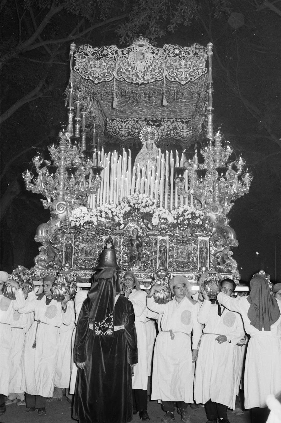 El trono de la Virgen de la Esperanza, enorme barco barroco donde los haya, surca la Alameda Principal en 1959. La grandiosidad de la obra que dirigió Adrián Risueño contrasta con la sencillez del atavío de la imagen, con una mantilla sobre la cabeza y una corona de casco completada con una ráfaga trasera circular.