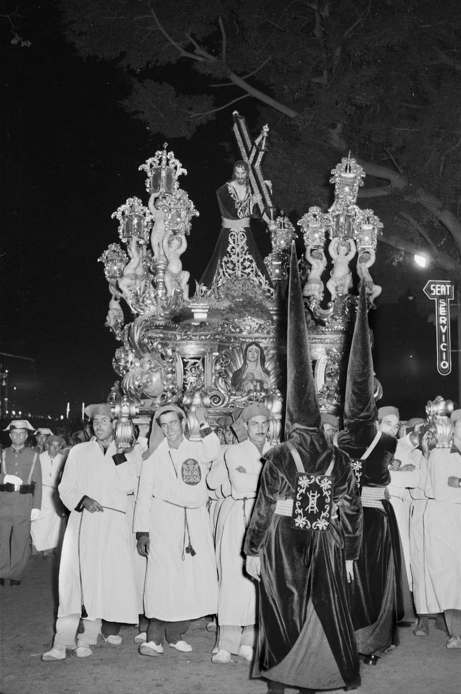 Jueves Santo de 1959. El Nazareno del Paso recorre la Alameda Principal en el trono que diseñara Fernando Guerrero-Strachan y ejecutara Francisco Palma Burgos entre 1940 y 1942. Todavía luce unos arbotantes configurados por brazos sobre los que unas figuras infantiles sostienen faroles. Posteriormente, fueron suprimidos los brazos para que esta especie de querubines descansaran directamente sobre el cajillo.