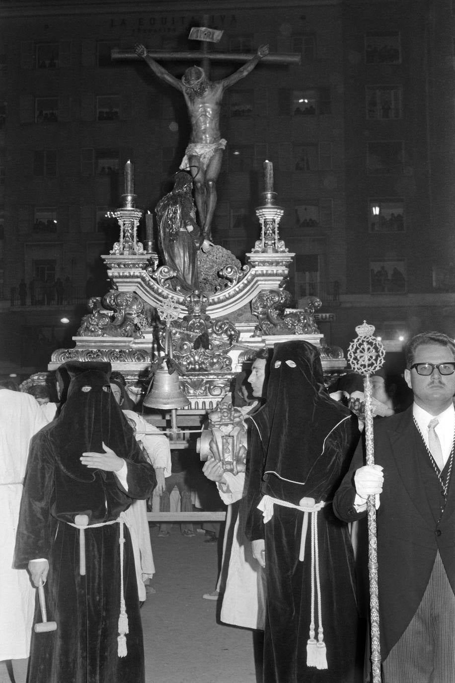 En esta fotografía de 19872, observamos el trono del Cristo de la Buena Muerte detenido en la rotonda del Marqués de Larios ante un habitado edificio de La Equitativa, hoy destinado a uso hostelero. Llama la atención lo reducido del tamaño de los cirios de las esquinas del trono.