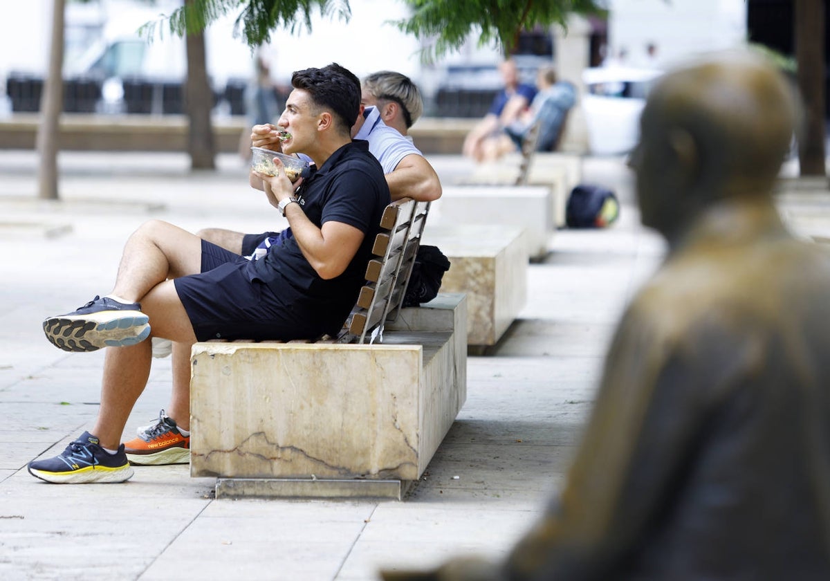 Un turista se come una ensalada en un banco de la plaza de la Merced ante la 'mirada' de Picasso.