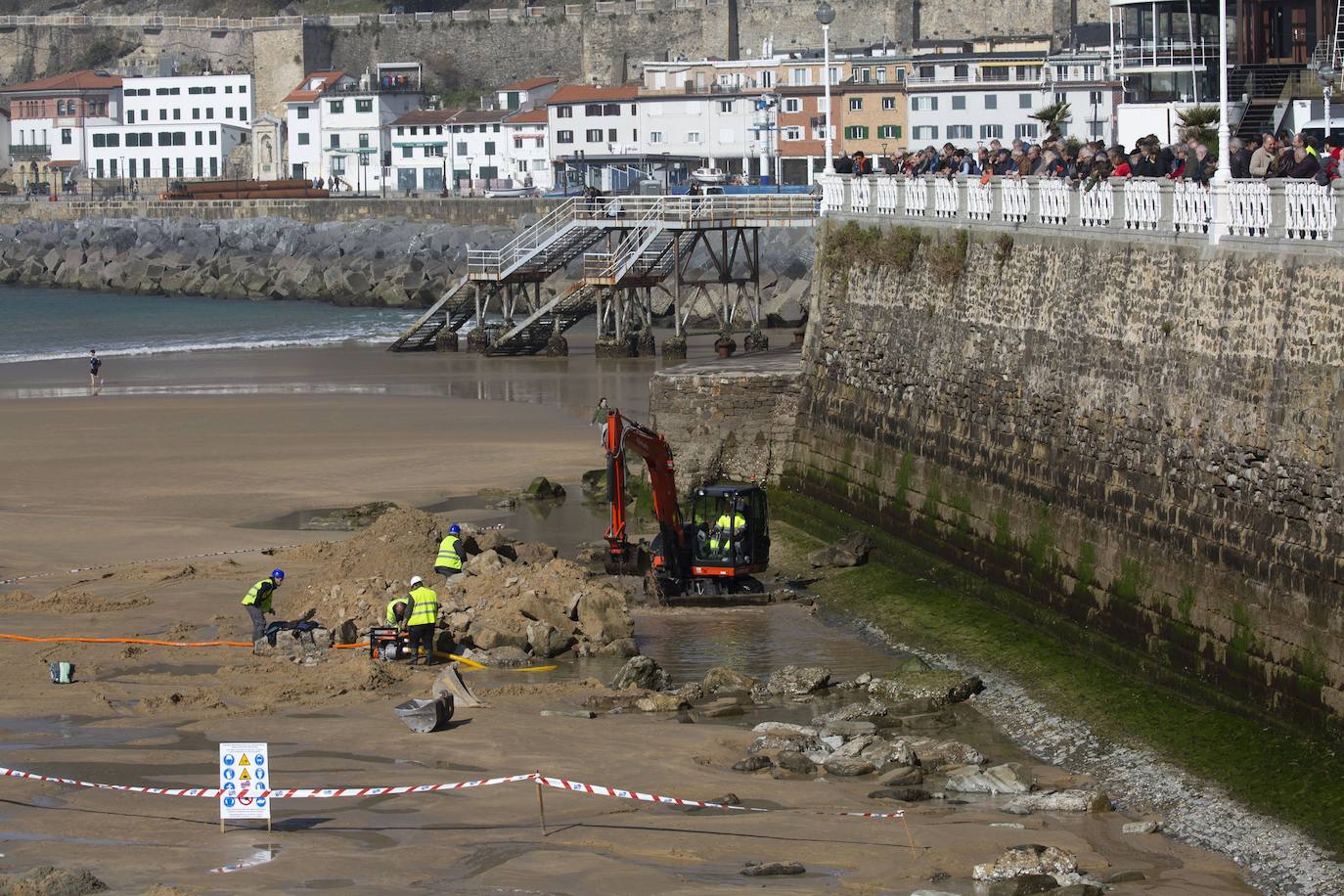 La playa de La Concha se ha convertido en la bajamar de este lunes por la mañana en un inesperado yacimiento arqueológico. La presencia de una pequeña excavadora retirando piedras y escombros junto al muro de costa a la altura del edificio consistorial ha suscitado una gran expectación entre los numerosos viandantes y turistas que han provechado el buen tiempo para dar un paseo. La máquina profundizaba en la arena en busca de los restos de un barco hundido del que hay constancia documental desde el siglo XVIII. 