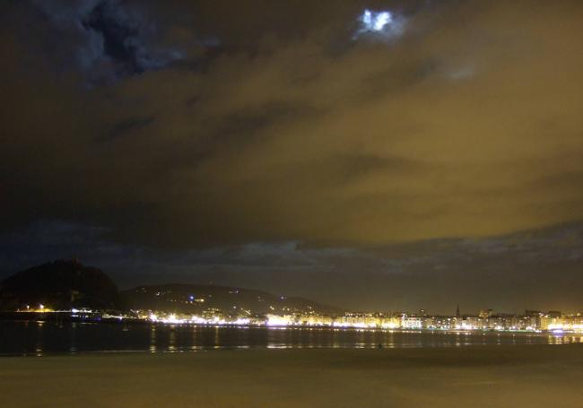 Las nubes de fondo se ven iluminadas en luz blanca grisácea por la Luna, mientras que en la zona central y superior derecha están iluminadas por la contaminación lumínica de San Sebastián.