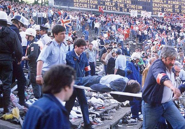Policías, sanitarios y voluntarios retiran a las víctimas, con los restos del desastre a la vista y los seguidores del Liverpool en la grada.