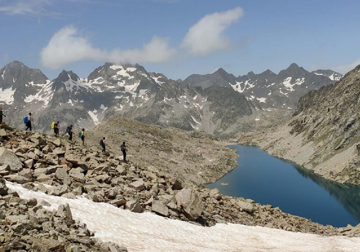Los montañeros avanzan entre neveros y rocas rumbo al Pico Serrato, con el Ibón de Bachimaña y las cumbres del macizo de los Infiernos al fondo.