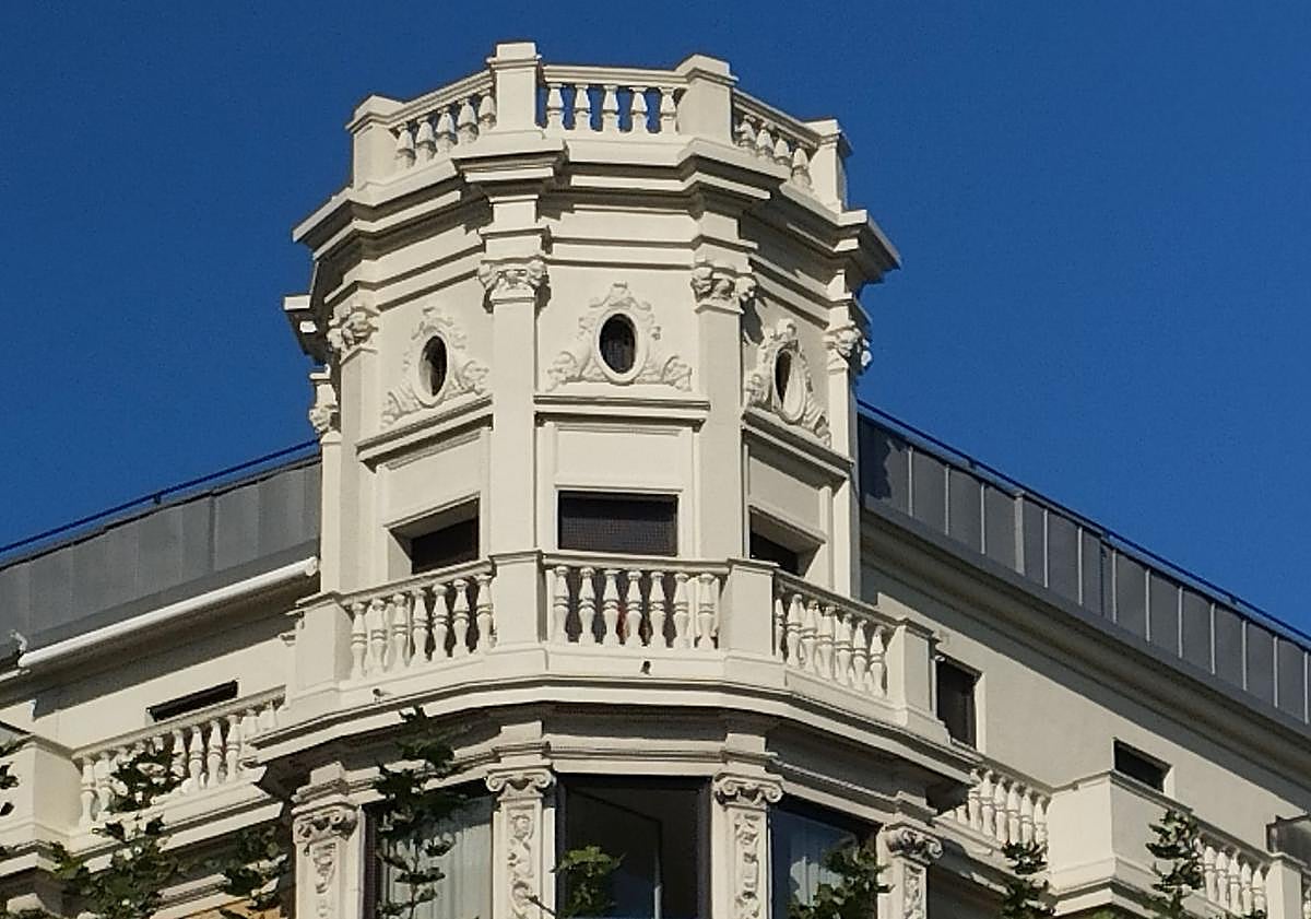 Imagen principal - Torre octogonal en el chaflán, con óculos, balaustrada y cornisas molduradas. El mirador de hormigón armado funciona como bisagra entre atmósferas urbanas. Debajo, balcones curvos en el centro de la fachada a Okendo, una unidad compositiva que agrupa tres alturas y ordena el alzado desde dentro.