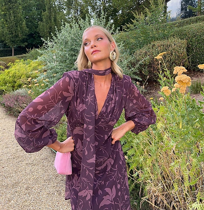 Vestido de estampado floral en morado, mangas abullonadas y escote en pico con bolso rosa.
