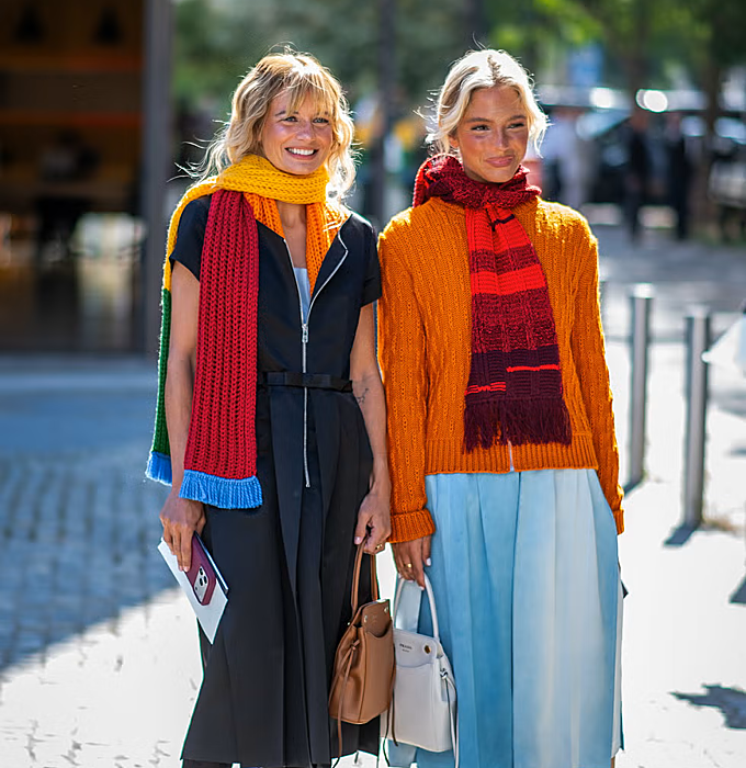Bufandas de punto en tonos vivos, jersey naranja de trenzas y falda plisada azul cielo.