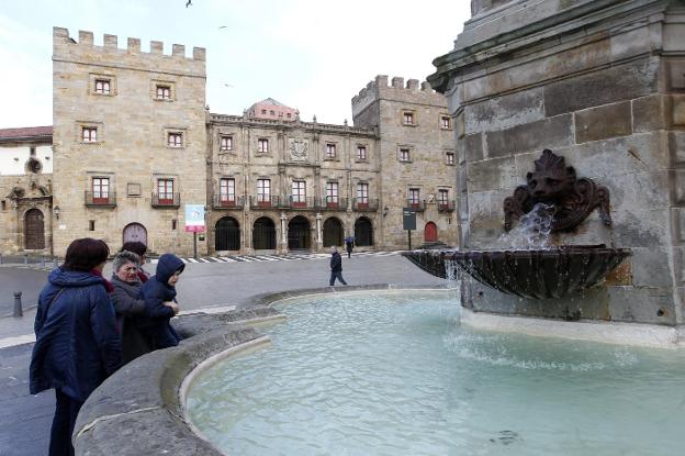 Varias personas contemplan la fuente de la estatua de Pelayo con el Palacio de Revillagigedo al fondo. 