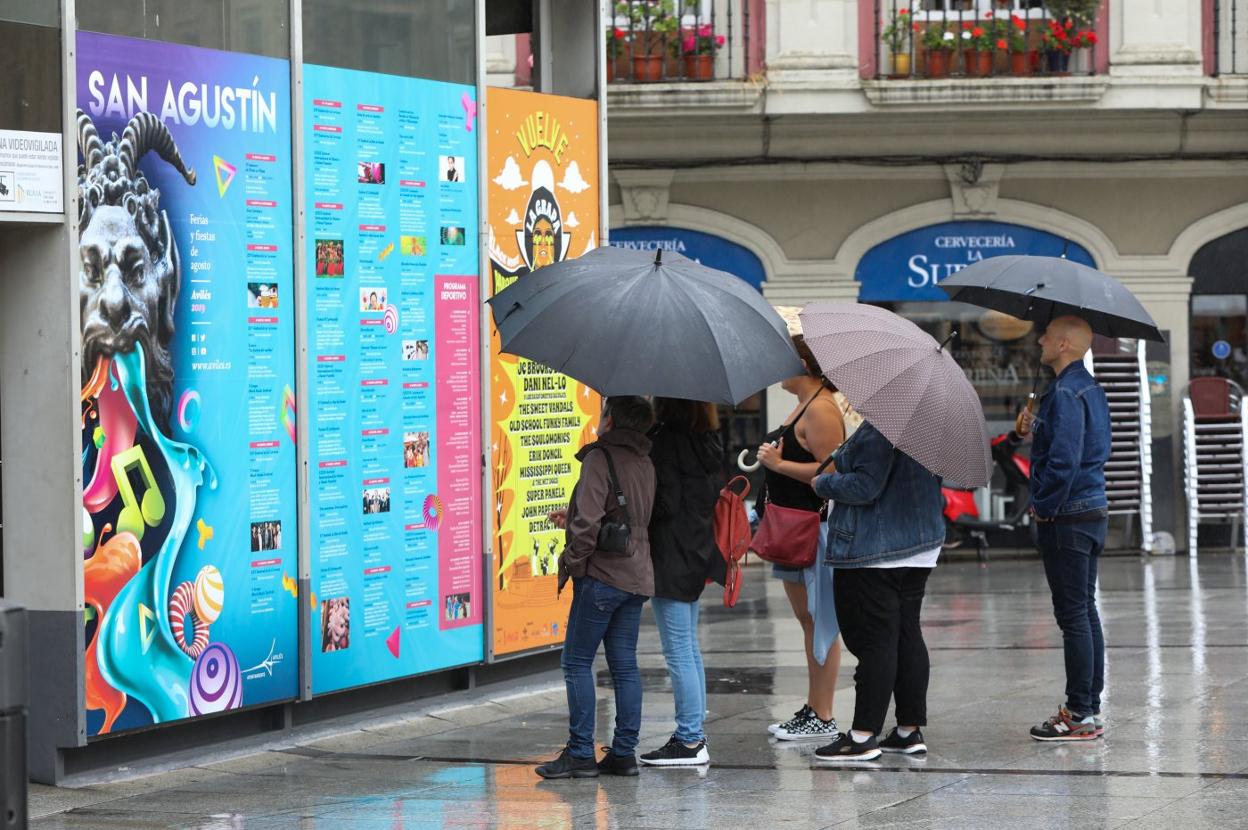 Un grupo observa el panel de la programación festiva de agosto protegidos de la lluvia con paraguas. 