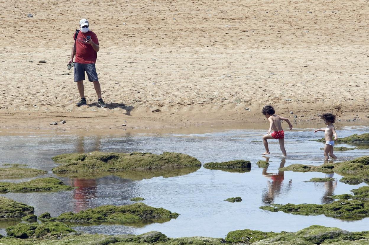 Unos niños juegan en el agua de la playa de San Lorenzo ante la mirada de su padre. 