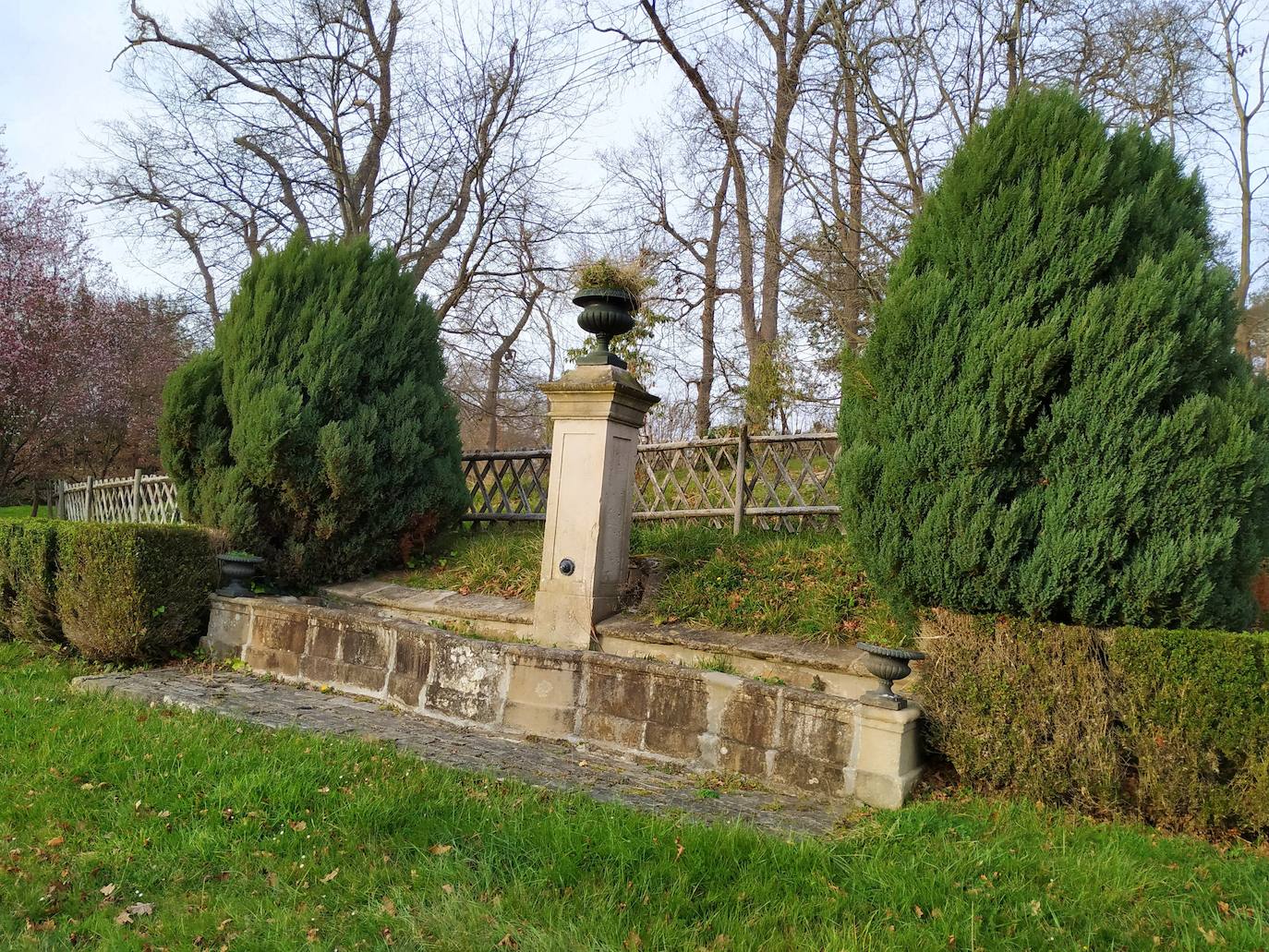 Fuente mural de piedra rematada con una copa del siglo XIX y flanqueada por dos enebros chino (Juniperus chinensis) situada entre el jardín en espiral y la trasera de la residencia de Álvaro Armada.