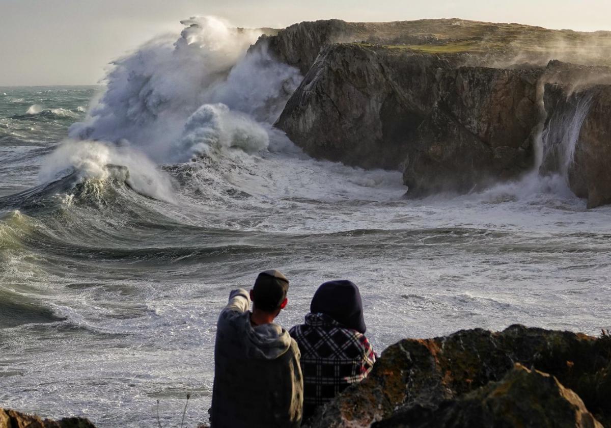 Dos personas observan el oleaje en la costa llanisca, junto a los acantilados de Pría, que superan los 20 metros de altura, pese a lo cual las olas llegan a barrer la senda costera.