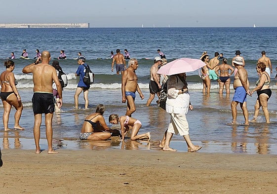 Bañistas disfrutando de una jornada estival en la playa de San Lorenzo, por la que una persona pasea con un paraguas para protegerse del sol.