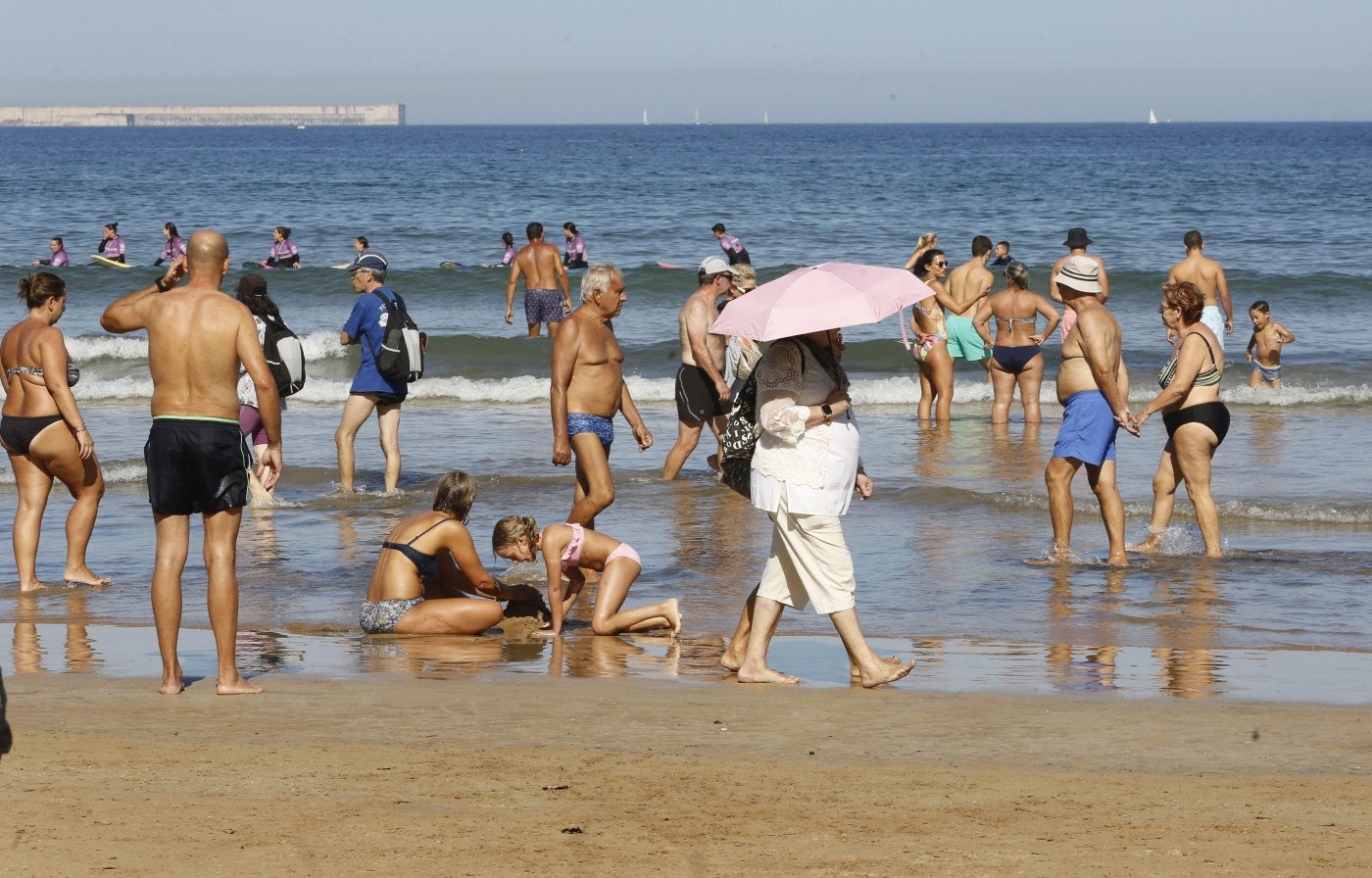 Bañistas disfrutando de una jornada estival en la playa de San Lorenzo, por la que una persona pasea con un paraguas para protegerse del sol.