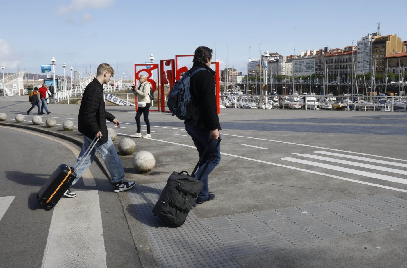 Dos turistas llegan con sus maletas a la ciudad y pasan frente a 'Las letronas', en el Puerto Deportivo.