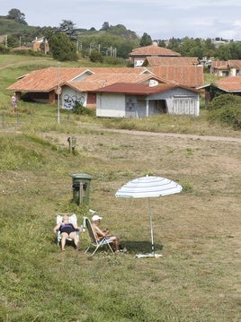 Dos personas toman el sol, en Perlora, con los edificios de la ciudad vacacional, en mal estado, al fondo.