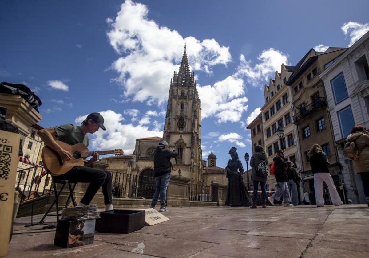 Un joven toca una guitarra en la plaza de la catedral de Oviedo mientras los turistas hacen fotos de la escultura de La Regenta