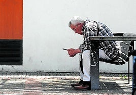 Un jubilado observa su móvil sentado en un banco a la sombra.