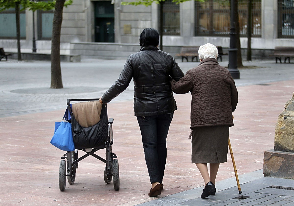 Una mujer, usuaria de silla de ruedas, pasea apoyada en un bastón y del brazo de su cuidadora.
