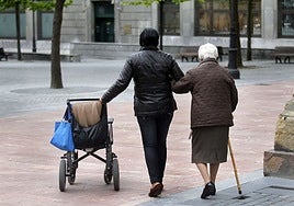 Una mujer, usuaria de silla de ruedas, pasea apoyada en un bastón y del brazo de su cuidadora.