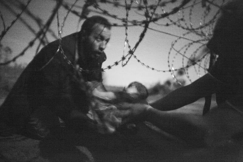 La fotografía, en blanco y negro, muestra un hombre pasando a un bebe a través de la valla de alambre de espino en Roeszke, en la frontera entre Serbia y Hungría, el 28 de agosto de 2015. Ganadora absoluta del World Pres Photo.