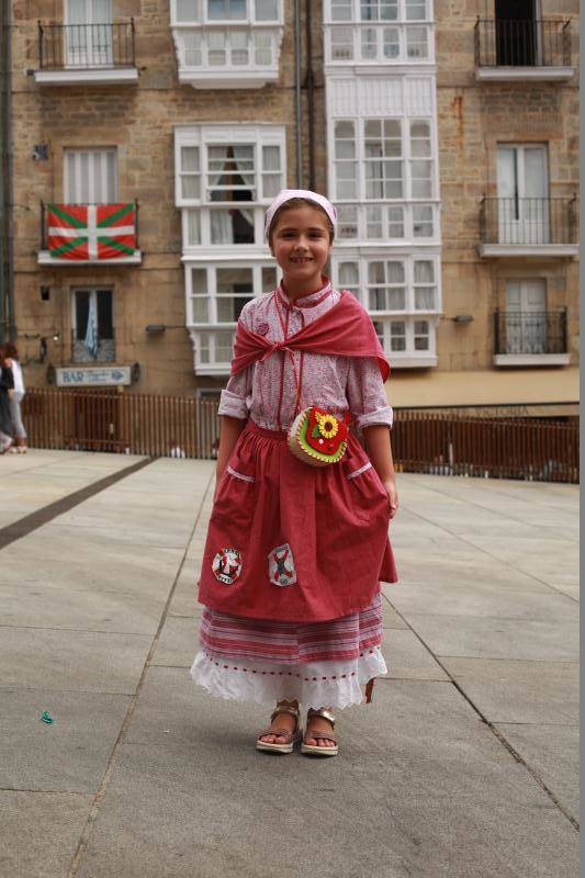 La pequeña Ane Martínez de Olcoz luce un traje rosa con flores y falda de rayas y como complemento un colorido bolso repleto de pegatinas de cuadrillas de blusas.