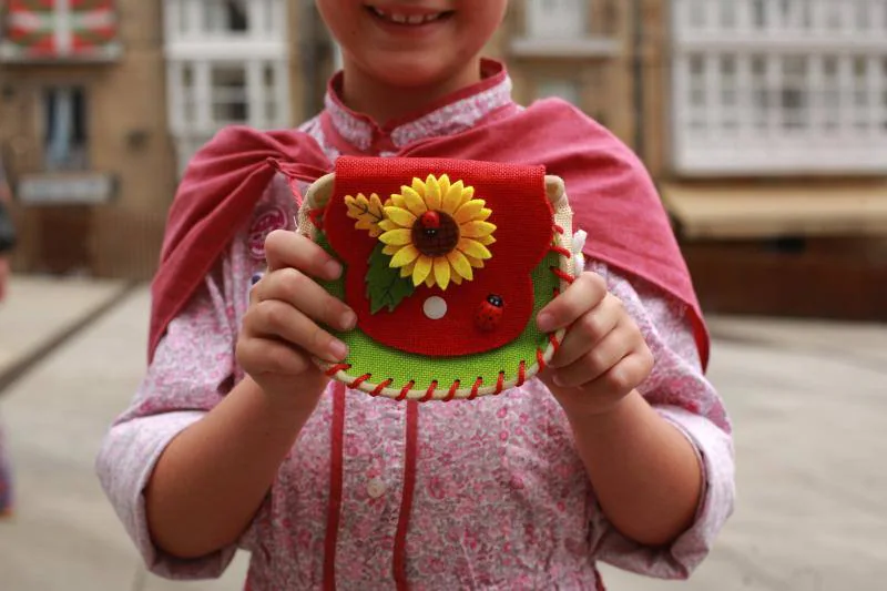 La pequeña Ane Martínez de Olcoz luce un traje rosa con flores y falda de rayas y como complemento un colorido bolso repleto de pegatinas de cuadrillas de blusas.