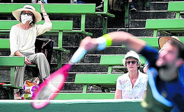 Imagen principal - Torneo tóxico. Una mujer con mascarilla sigue un partido clasificatorio en Melbourne. Abajo, el humo sobre la pista central y un miembro de la organización se protege del aire.