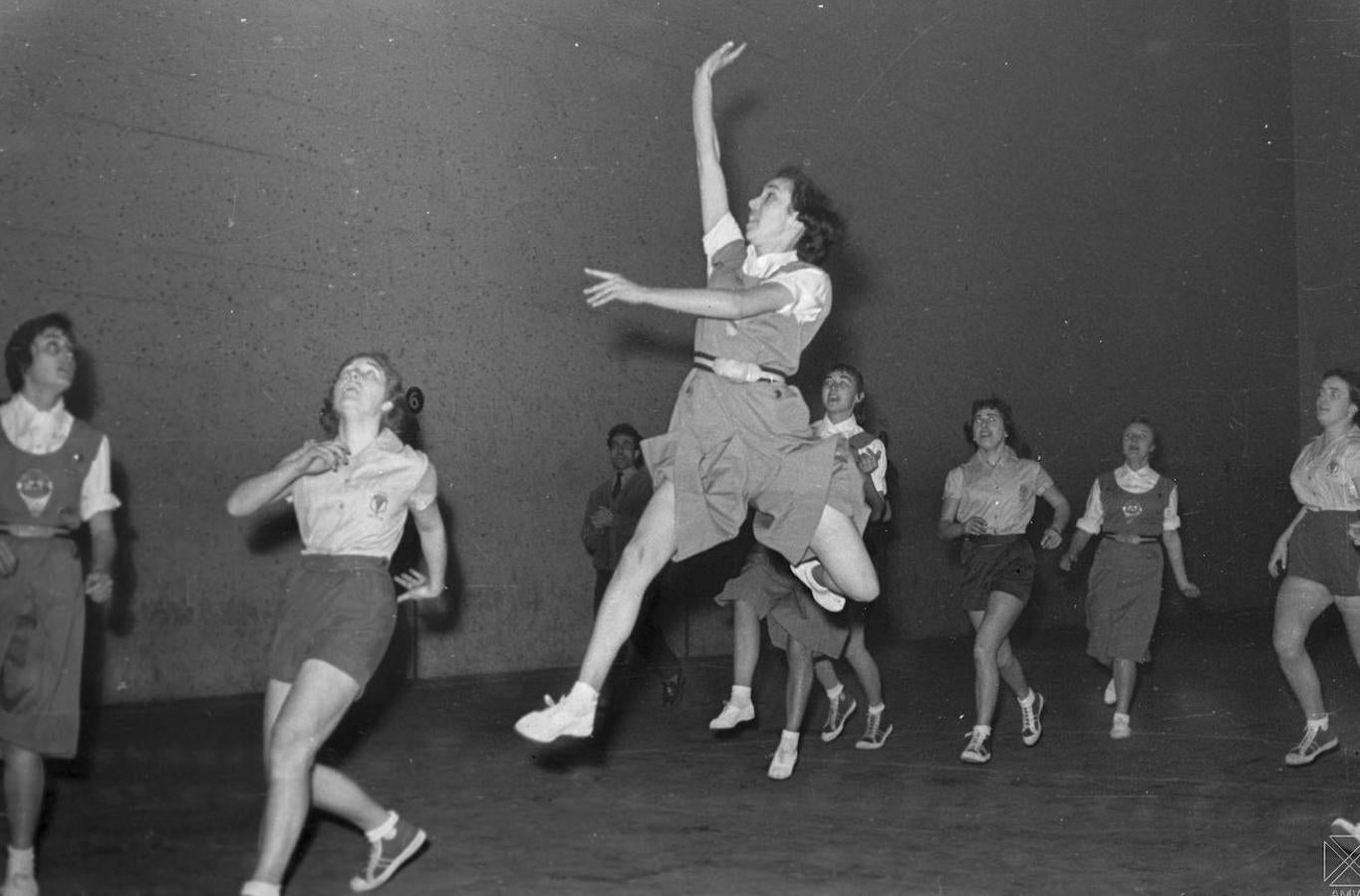 Aunque se sale del marco de la imagen captada por Fede Arocena, el balón participó del juego. Así lo atestigua tanto el estiloso lanzamiento a canasta de la joven vestida con falda deportiva, perteneciente al equipo de Álava, como el resultado, un raquítico 22-18 a favor del Imosa. El partido amistoso se disputó el 22 de enero de 1958 en el frontón Vitoriano, que de vez en cuando hacía un hueco dentro de su amplia oferta pelotazale para ofrecerse como cancha del baloncesto alavés. En esas fechas, el básquet femenino local pasaba por un mal momento después de años boyantes, con solo dos conjuntos en acción. Uno estaba vinculado a la fábrica DKW y lo había promovido una trabajadora alemana que, además, trajo el pantalón corto que se aprecia en la foto y el otro, el Álava, impulsado por la propia federación para agrupar a las jugadoras que se habían quedado sin equipo por la desaparición del suyo. Tampoco el campeonato provincial masculino gozaba de buena salud en 1958. Había una competición reglada que por lo general disputaba sus jornadas en el campo militar de Flandes, pero la poca seriedad de los contendientes se manifestaba a veces por las incomparecencias de ellos. Jugaban aquella liga el Vitoria, El Pilar, San José, Helios, Juvalde, Areitio y San Fernando.