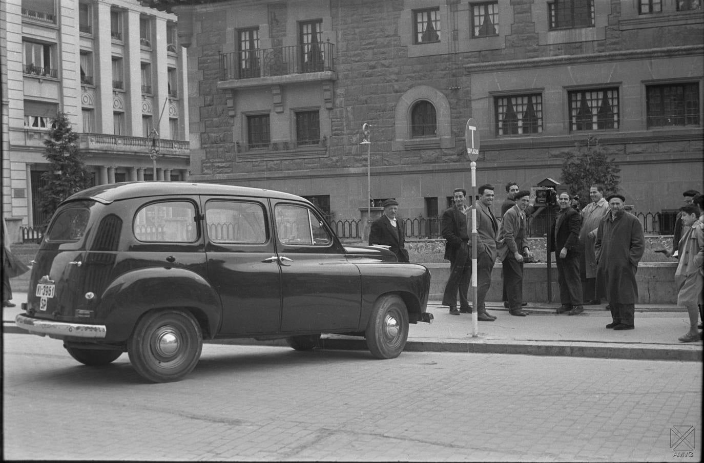 Los más de veinte vehículos que disponían de licencia de taxi en Vitoria en 1957 estrenaron taxímetro el 24 de marzo tras ser verificados por el Ayuntamiento. El concejal delegado de Circulación era el médico José María Viana, con quien los conductores acordaron también la instalación de una luz verde (chivato) en el coche para advertir al cliente de que estaba libre. Durante el día, la disponibilidad del servicio la marcaba la posición de la bandera del nuevo dispositivo, subida (vacío) o bajada. Las novedades en los taxis aconsejaron la actualización de las tarifas entre el Ayuntamiento y el gremio. Así, la bajada de bandera se estableció en cinco pesetas, el kilómetro en cuatro y el suplemento por salir al extrarradio se incrementaba en 3, 6 y 9 si eran uno, dos o tres los kilómetros más allá de los límites. Por taxi solo se aceptaban tres viajeros y si se superaban, había que abonar dos pesetas por cada uno de más. También se tarifaron los servicios especiales: boda antes de las 11 de la mañana (100 pesetas), después (125), bautizo en el casco urbano (50), conducción (75) y a la plaza de toros y al campo de fútbol (15). Por la noche, los precios se doblaban. Los taxistas vitorianos estrenaron por entonces la parada principal, al inicio de la calle Olaguíbel, detrás de Correos, que disponía de teléfono y urinarios, aunque no de sombra, como muestra la foto. Hasta que creció el arbolado allí plantado, se protegían con una sombrilla cuando daba el sol. Tuvieron también sus más y sus menos con el alcalde Luis Ibarra, que en diciembre de aquel año multó a trece de ellos con 50 ptas. por saltarse el horario de espera que les correspondía en la parada.