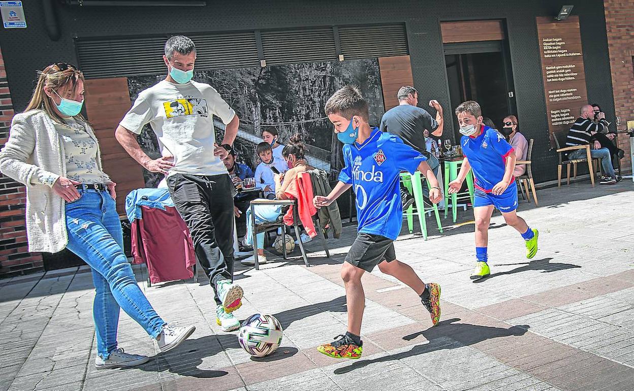 Dos chavales, con la camiseta de 'los azules', pasan corriendo en busca del balón frente a una terraza repleta. 