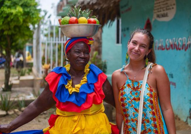 Ángela sonríe junto a una mujer palenquera, que con su alegría y ropa colorida ofrece exóticos frutos a los transeúntes en San Basilio de Palenque, primer pueblo negro libre de América.