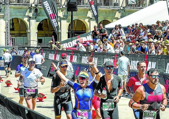 Triatletas, entre ellos una fémina que alza los brazos, pasan por la meta de la plaza de España en una de las vueltas de la prueba de a pie en el Ironman de Vitoria de 2022.