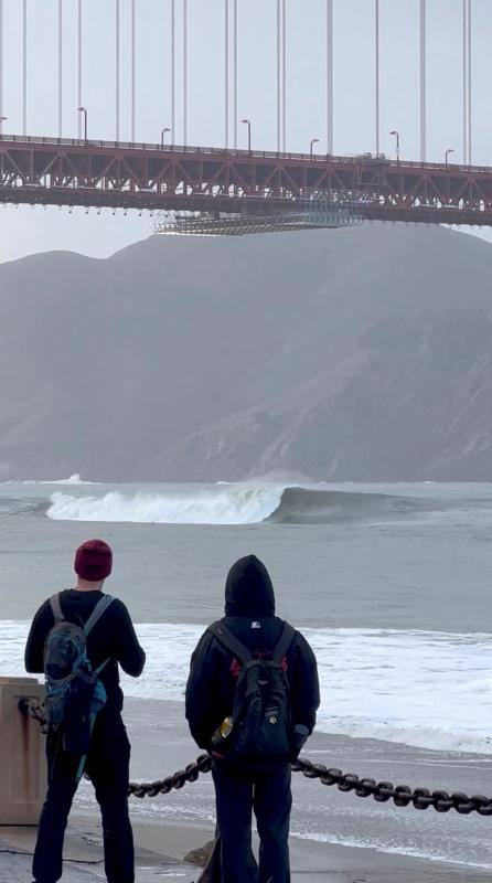 Dos jóvenes observan una imponente ola junto al emblemático puente de San Francisco.