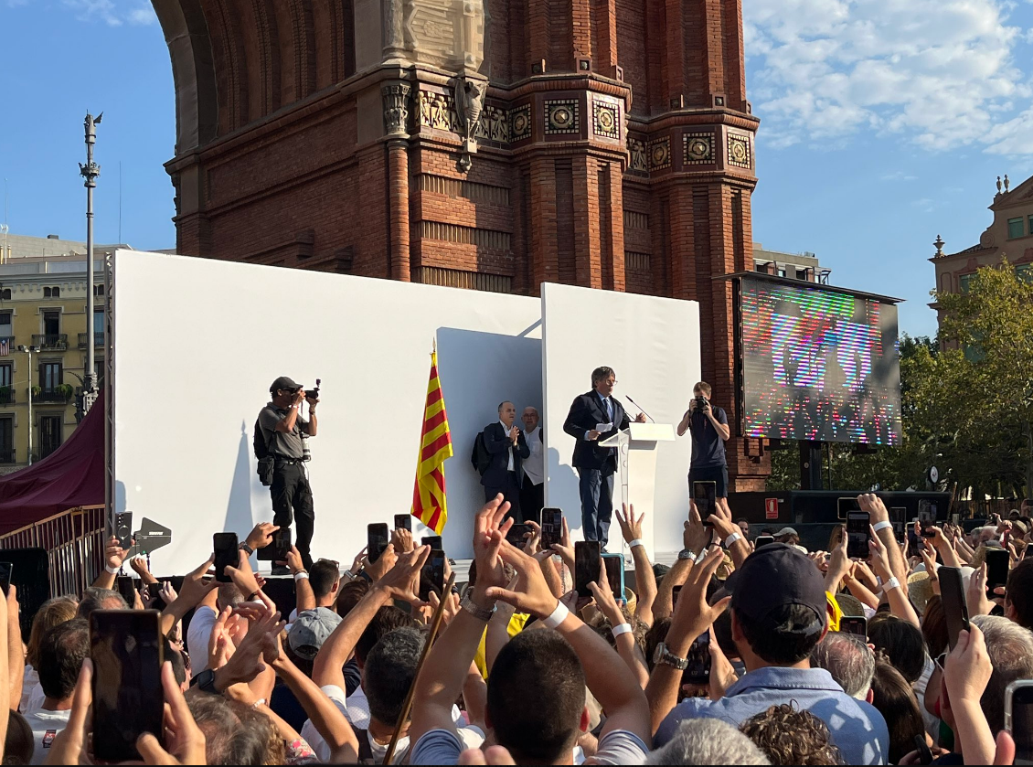Imagen del escenario en el que se ve a Gonzalo Boye, vestido con camisa blanca, esperando a que Puigdemont finalice su discurso y ayudarle a huir.