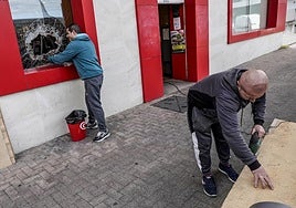 Un responsable de la tienda retira esquirlas de cristal y otro prepara un tablón de aglomerado para tapar el agujero.