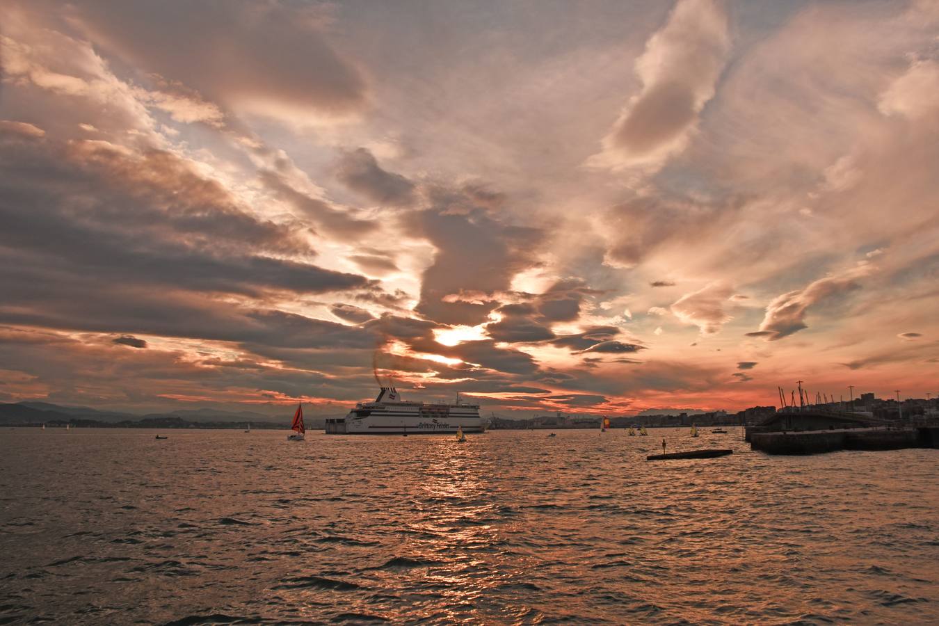 Un cielo parcialmente cubierto y teñido de rojo al atarceder vigila la bahía santanderina y la entrada del ferry.