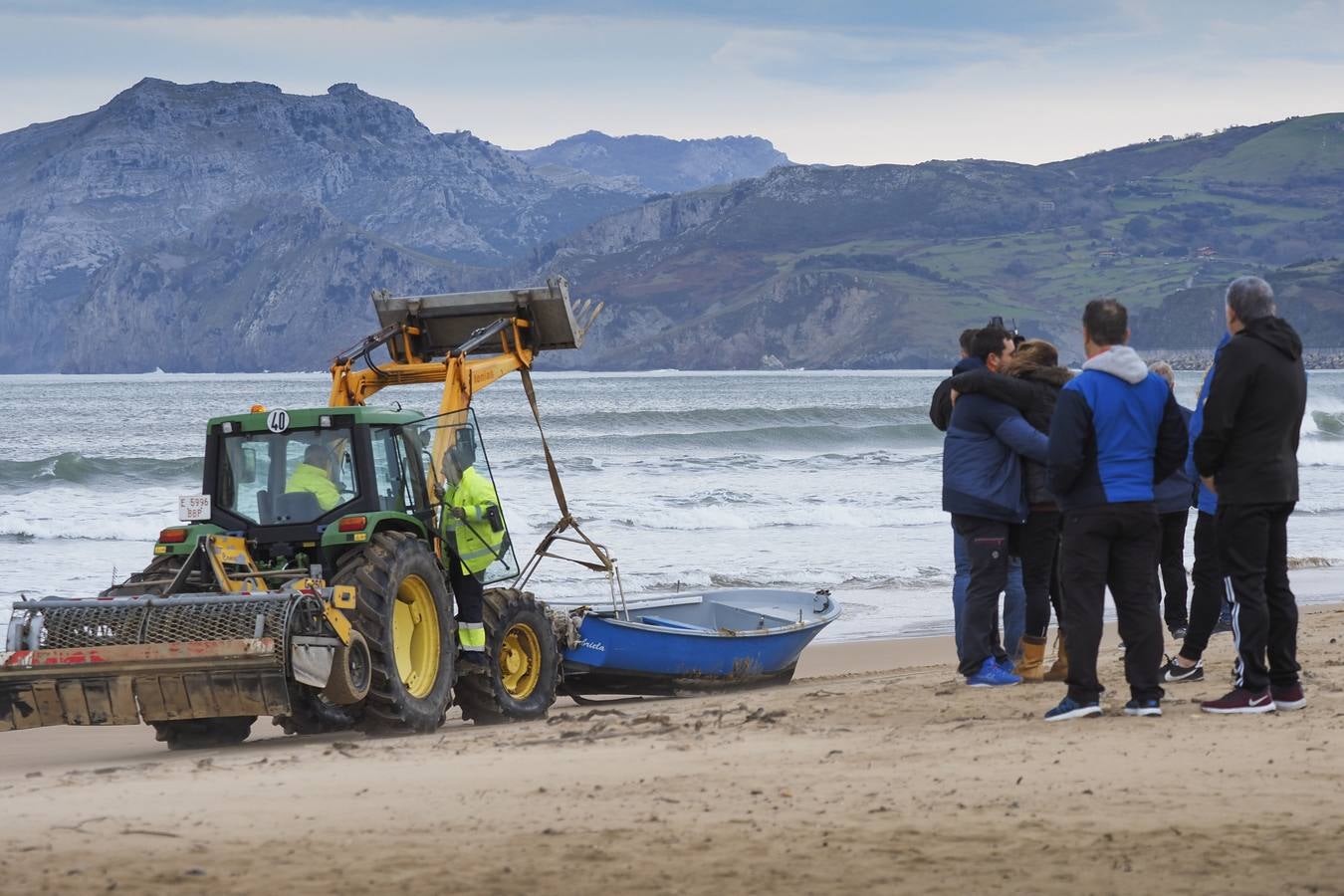 Familiares y amigos del pescador fallecido se abrazan en la playa Salvé a la espera de noticias, mientras un tractor remolca la embarcación siniestrada.