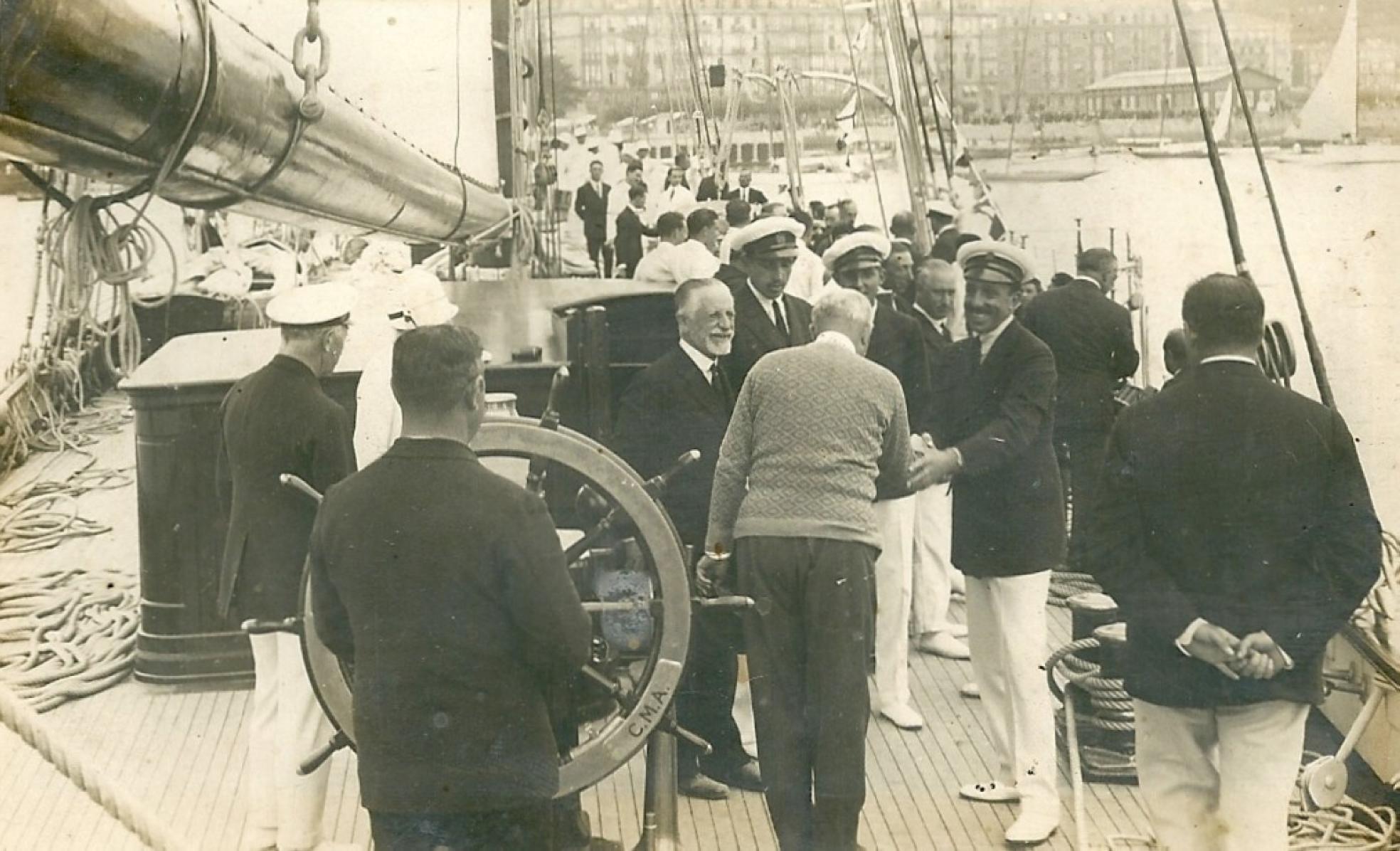 Alfonso XIII felicita a Horacio Echevarrieta sobre la cubierta del María del Carmen Ana, en presencia del capitán Ramón Urrechaga (al timón) y de los infantes don Jaime y don Juan, entre otras personalidades, tras la finalización en Santander de la regata entre Plymouth y la capital cántabra el 29 de agosto de 1929 en la que se impuso. 