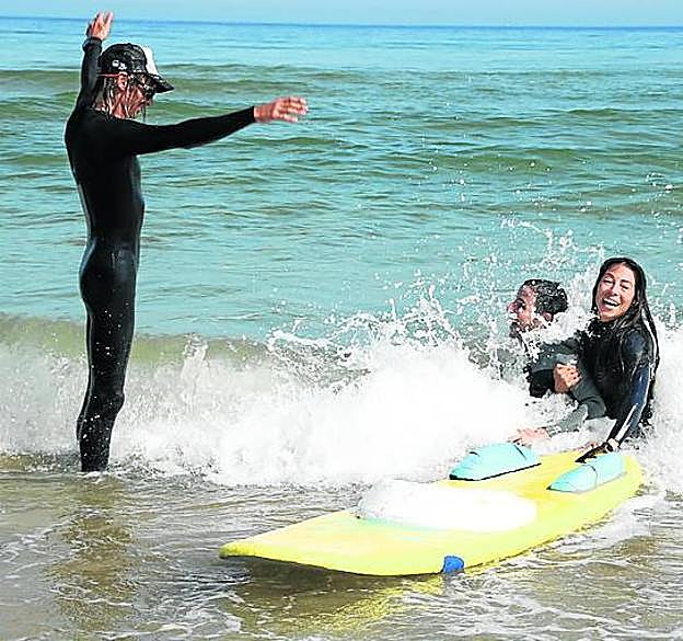 Nancy Rojas ayuda a salir del mar a Héctor Salgado mientras les empuja una ola y Thomas Heller, de pie, se divierte con la escena.