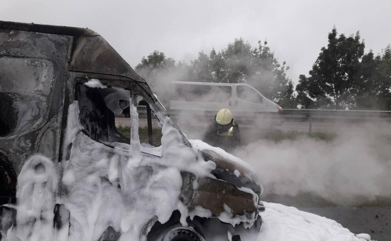 Un bomberos sofoca las llamas de la furgoneta, que ha quedado totalmente calcinada tras incendiarse cuando circulaba por la carretera de Liérganes